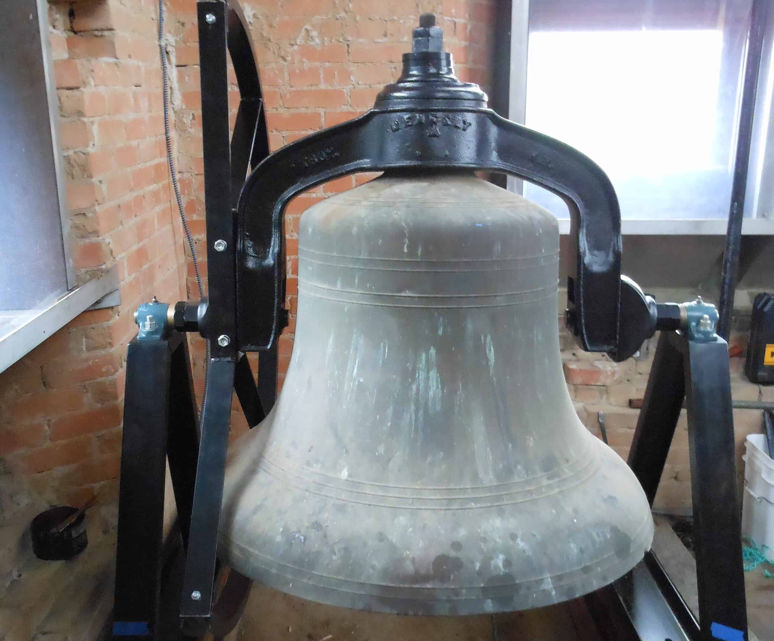 Bell at Saint Andrew's Episcopal Church on display, mounted on a black metal frame inside a brick-wa.
